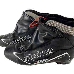 Peut inclure: Paire de chaussures de ski de randonnée Alpina TS Plus noires. Les chaussures ont un extérieur noir avec des accents blancs et rouges, et le nom de la marque est bien visible. Les chaussures ont une conception montante avec un col en mesh et des lacets rouges.