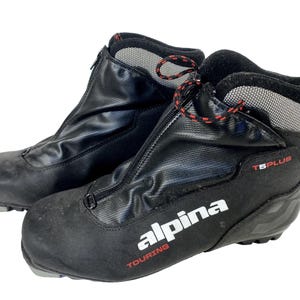 Peut inclure: Paire de chaussures de ski Alpina Touring noires. Les chaussures ont une fermeture éclair sur le devant, des lacets rouges et noirs, et la marque "alpina" en blanc avec "TOURING" en rouge. Les bottes sont étiquetées "T6 PLUS".