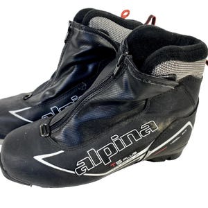 Peut inclure: Paire de chaussures de ski Alpina TS Plus noires. Les bottes ont une fermeture éclair sur le devant, des accents blancs et des tirettes rouges. Le nom de la marque est affiché en blanc. Pour les sports d'hiver.