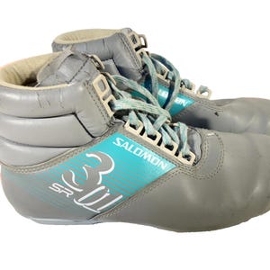 Puede incluir: Par de botas de esquí Salomon SR grises con detalles y cordones turquesa. Las botas tienen un diseño de caña alta con hebillas blancas y el logotipo de Salomon. Diseñadas para esquí alpino.