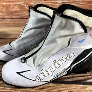 Peut inclure: Paire de chaussures de ski de fond blanches et grises avec des accents noirs. Les bottes présentent la marque "Alpina" et le texte "TS Plus Touring". Les bottes ont une fermeture éclair et un ajustement de talon noir.