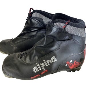 Peut inclure: Paire de chaussures de ski de randonnée Alpina T5 JR Plus noires pour juniors. Les chaussures ont une fermeture éclair, des détails rouges et le logo Alpina en blanc. Conçues pour les jeunes skieurs.