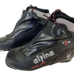 Peut inclure: Paire de chaussures de ski de randonnée Alpina T5 JR Plus noires. Les chaussures ont une fermeture éclair sur le devant, des lacets rouges et noirs et le logo Alpina en blanc. Conçues pour les jeunes skieurs.