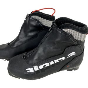 Peut inclure: Paire de chaussures de ski de fond noires. Les chaussures ont une fermeture éclair sur le devant, une boucle de tirage rouge et du texte blanc sur le côté. Les bottes sont conçues pour les sports d'hiver et les activités de plein air.