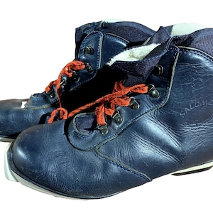 Puede incluir: Par de botas de esquí Salomon azul marino vintage con cordones naranjas. Las botas tienen un diseño de caña alta con ojales metálicos y una suela blanca. El logotipo de Salomon es visible en el lateral.