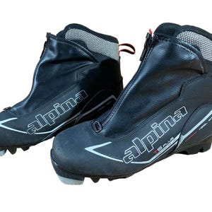 Peut inclure: Paire de chaussures de ski de fond Alpina TS Plus noires. Les bottes ont une fermeture éclair sur le devant, des accents blancs et rouges, et le logo Alpina en blanc. Les bottes sont conçues pour les sports d'hiver.