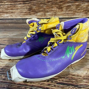 Puede incluir: Par de botas de esquí de fondo Salomon vintage moradas y amarillas. Las botas tienen cordones y detalles amarillos, con gráficos verdes y amarillos. Las botas están sobre una superficie de madera.