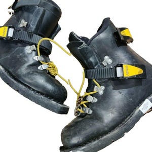 May include: Pair of black leather mountaineering boots with yellow accents. The boots feature metal hardware, yellow laces, and adjustable straps with yellow buckles. The soles are thick and rugged, designed for challenging terrain.