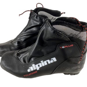 Peut inclure: Paire de chaussures de ski de randonnée Alpina T5 Plus noires. Les chaussures ont une fermeture éclair sur le devant, des lacets rouges et noirs et le logo Alpina en blanc. Conçues pour les sports d'hiver et les activités de plein air.