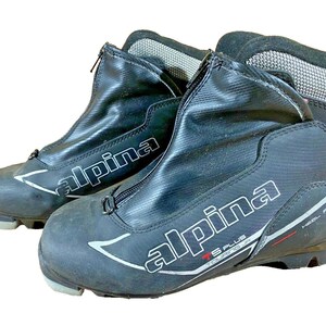 Peut inclure: Paire de chaussures de ski de fond Alpina noires. Les bottes ont une fermeture éclair sur le devant, des accents blancs et rouges et le logo Alpina. Les bottes sont conçues pour le touring et ont une fonction de maintien du talon.