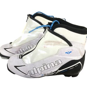 Peut inclure: Paire de chaussures de ski de fond Alpina TS Plus blanches et noires. Les chaussures ont une tige blanche avec des accents noirs, des lacets bleus et le logo Alpina. Conçues pour le ski de fond.