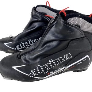 Peut inclure: Paire de chaussures de ski de fond Alpina noires. Les chaussures ont une fermeture éclair sur le devant, la marque Alpina en blanc et des accents rouges. Les chaussures sont conçues pour le ski de fond et présentent un design HEEL FIT.