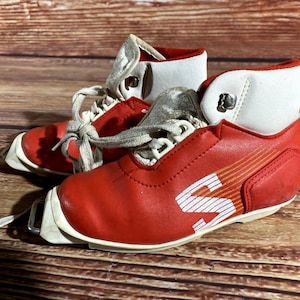 Puede incluir: Un par de botas de esquí rojas y blancas vintage. Las botas tienen cordones blancos, un puño blanco y la letra "S" en blanco en el lateral. Las botas son de cuero y están sobre una superficie de madera.