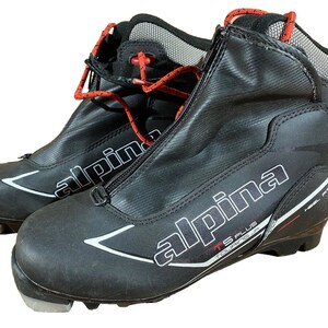 Peut inclure: Paire de chaussures de ski de randonnée Alpina TS Plus noires. Les chaussures ont une fermeture éclair sur le devant, des lacets rouges et des accents blancs et rouges. Conçues pour les sports d'hiver.
