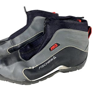 Puede incluir: Par de botas de esquí de fondo K2 Comfort Fischer negras, grises y rojas. Las botas tienen una cremallera frontal, una parte superior de tela negra y una superposición de cuero sintético gris. El logotipo de Fischer es visible en el lateral.