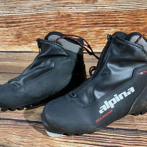 Peut inclure: Paire de chaussures de ski de randonnée Alpina T6 Plus noires. Les chaussures ont une fermeture éclair avant, des accents rouges et gris, et le logo Alpina en blanc. Les chaussures sont sur une surface en bois.