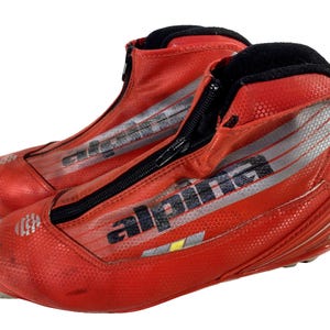 Puede incluir: Un par de botas de esquí rojas con cremalleras negras y el nombre de la marca "Alpha" impreso en el lateral. Las botas tienen una superficie texturizada y un forro negro en la parte superior. Diseñadas para deportes de invierno.