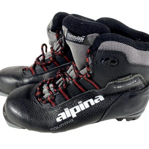 Peut inclure: Paire de chaussures de ski de randonnée Alpina noires avec des lacets rouges et noirs. Les chaussures ont un extérieur en cuir texturé, des accents en tissu gris et noir et le logo Thinsulate Insulation.