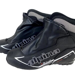Peut inclure: Paire de chaussures de ski de fond noires avec le nom de marque "alpina" en blanc. Les chaussures ont une fermeture éclair sur le devant, un col en mesh et un accent rouge. Elles sont conçues pour un ajustement au talon, sur fond blanc.
