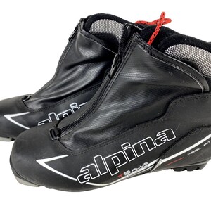Peut inclure: Paire de chaussures de ski de fond noires avec la marque "Alpina" en blanc. Les bottes ont une fermeture éclair sur le devant, un détail de lacet rouge et un motif de flèche blanche. Les bottes sont conçues pour un ajustement au talon.