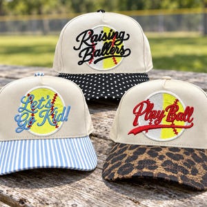 May include: Three baseball caps with cream-colored crowns and embroidered softball graphics. The top cap has a black and white polka dot brim and says Raising Ballers. The left cap has a blue and white striped brim and says Let's Go Kid!. The right cap has a leopard print brim and says Play Ball.