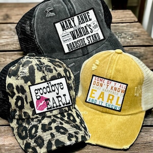 May include: Three distressed trucker hats with mesh backs. The black hat features a patch reading 'MARY ANNE and WANDA'S ROADSIDE STAND'. The leopard print hat says 'goodbye EARL' with a pink lip icon. The yellow hat says 'SOME OF Y'ALL DON'T KNOW WHAT HAPPENED TO EARL AND IT SHOWS'.