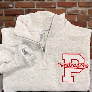 May include: A light gray quarter-zip sweatshirt with the word "Pendragons" embroidered in red. The sweatshirt has a folded sleeve and is displayed on a white wooden surface.
