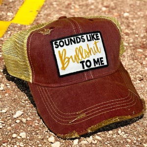 May include: A distressed maroon baseball cap with a gold mesh back. The front features a rectangular white patch with black text that reads SOUNDS LIKE and TO ME, with the word Bullshit written in gold cursive script in the center.