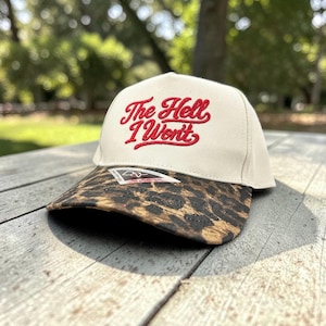 May include: A cream-colored baseball cap featuring the red embroidered text The Hell I Won't on the front panel. The brim of the cap displays a brown and black leopard print pattern and includes a small rectangular product tag attached to the center.