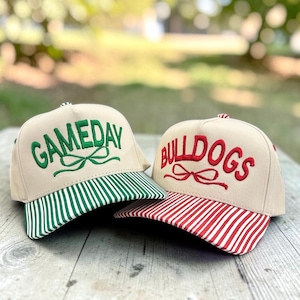 May include: Two beige baseball caps with embroidered text and bows. The left cap features green text reading GAMEDAY with a green bow and a green-and-white striped brim. The right cap features red text reading BULLDOGS with a red bow and a red-and-white striped brim.