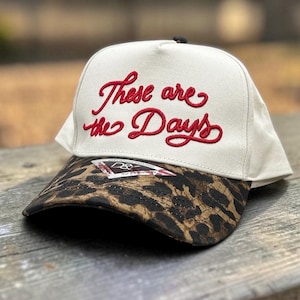 May include: A cream-colored baseball cap featuring the embroidered red script text These are the Days on the front panel. The curved brim displays a brown and black leopard print pattern, and a small triangular brand label is attached to the brim.