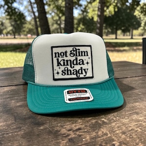 May include: A teal and white trucker hat with the text "not slim kinda shady" in a black square. The hat has a white front panel, a teal brim, and a green mesh back. The hat is sitting on a wooden surface.