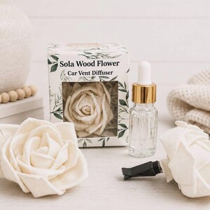 May include: A Sola Wood Flower Car Vent Diffuser set featuring a white box with green leaf illustrations, a glass bottle with a gold dropper cap, and two cream-colored wooden rose flowers, one attached to a black plastic car vent clip.
