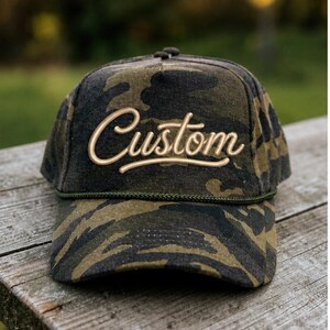 May include: A camouflage print baseball cap featuring the word Custom in raised, light-colored script lettering across the front panel. The hat includes a dark green braided cord trim along the base of the crown, just above the curved brim.