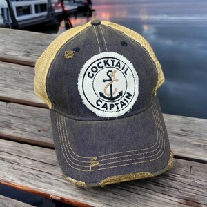 May include: A charcoal gray baseball cap with a distressed finish, featuring a tan mesh back and a circular white patch on the front. The patch displays a black anchor graphic and the text COCKTAIL CAPTAIN in bold black capital letters.