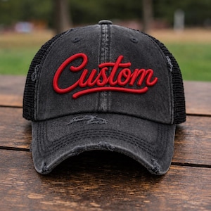 May include: A charcoal grey distressed baseball cap with a black mesh back and a curved brim. The front panel features the word Custom embroidered in a bold, raised red script font. The fabric has a faded, worn-in appearance with frayed edges.