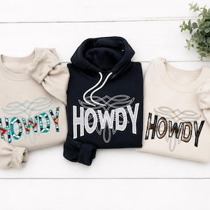 May include: Two beige crewneck sweatshirts and one black hooded sweatshirt, each featuring the word HOWDY in large capital letters with distinct patterns, including geometric shapes, white dots, and brown plaid, centered above a thin line graphic.