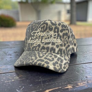 May include: A baseball cap featuring a black and tan leopard print pattern. The front of the cap displays the embroidered text "forever chasing weekends" in a cream-colored cursive script font, centered on the crown above the curved brim.