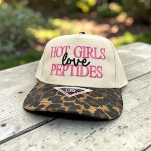 May include: A beige baseball cap with a leopard print brim. The front panel features embroidered text in pink and black that reads HOT GIRLS love PEPTIDES. A small triangular sticker with the text DAVID & YOUNG D&Y is attached to the brim.