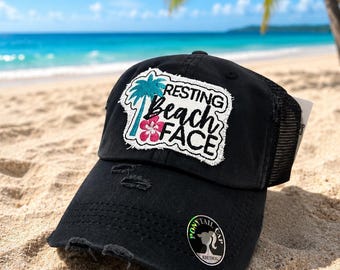 May include: A black distressed baseball cap with a mesh back and a white patch featuring a teal palm tree, a pink hibiscus flower, and the text Resting beach face. A circular sticker on the brim reads Pony tail cap Kbethos.