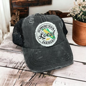 May include: A distressed charcoal gray baseball cap with a black mesh back. The front features a circular white patch with frayed edges, displaying a yellow corn cob graphic and the black text Support Your Local Farmers in a circular layout.