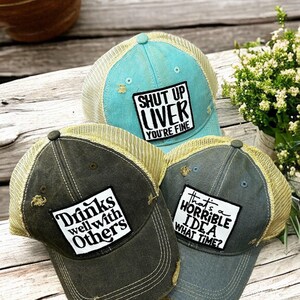 May include: Three distressed trucker hats with mesh backs. One is light blue with a patch reading SHUT UP LIVER YOU'RE FINE. One is dark gray with a patch reading Drinks well with Others. One is blue-gray with a patch reading that's a HORRIBLE IDEA WHAT TIME?
