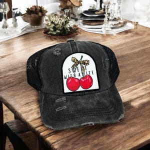 May include: Distressed black trucker hat with a mesh back. The front patch features two red cherries, a leopard print bow, and the text "Sweet with a Bite". The hat is sitting on a wooden table.