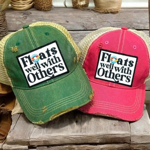May include: Two distressed trucker hats, one green and one red, with a tan mesh back. Each hat features a white square patch with the text "Floats well with Others" in black, and a small graphic.