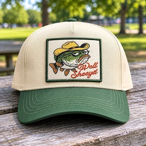 May include: A beige baseball cap with a dark green brim and a rectangular patch on the front. The patch features an embroidered green fish wearing a yellow cowboy hat and round glasses, with the orange cursive text Well Sheeyit below it.
