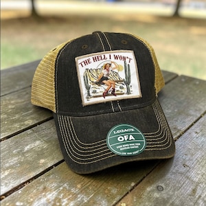 May include: A black and tan trucker hat with a patch that reads "THE HELL I WON'T" featuring a desert scene and a woman. The hat has a mesh back and a curved brim. The hat also has a green tag that says "LEGACY OFA".