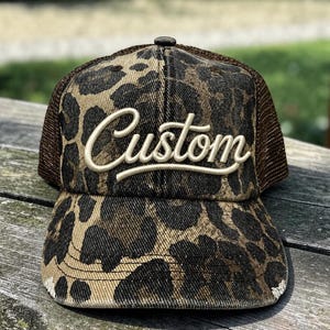May include: A trucker hat featuring a brown and black leopard print pattern on the front panels and brim. The word Custom is embroidered in a raised, cream-colored script font across the front. The back panels are made of brown mesh.