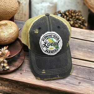 May include: A distressed black baseball cap with a khaki mesh back and a curved brim. The cap features a circular patch with the text "Support Your Local Farmers" and an image of corn. The cap has a small pin on the side.
