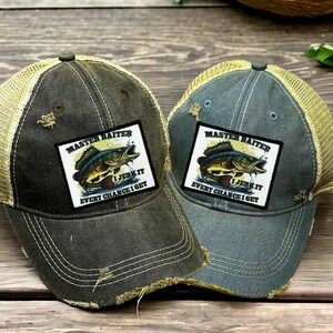 May include: Two distressed baseball caps with mesh backs, one dark gray and one blue-gray. Each features a rectangular patch with a graphic of a fish and the text MASTER BAITER I JERK IT EVERY CHANCE I GET. The caps have frayed edges on the brims.