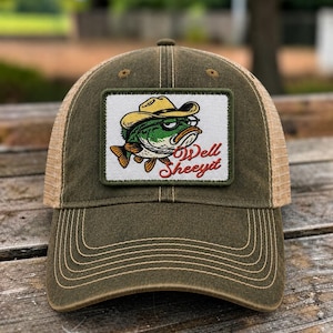 May include: An olive green baseball cap with a mesh back and a rectangular patch on the front. The patch features a cartoon fish wearing a yellow cowboy hat and glasses, with the red text "Well Sheeyit" printed below the fish illustration.
